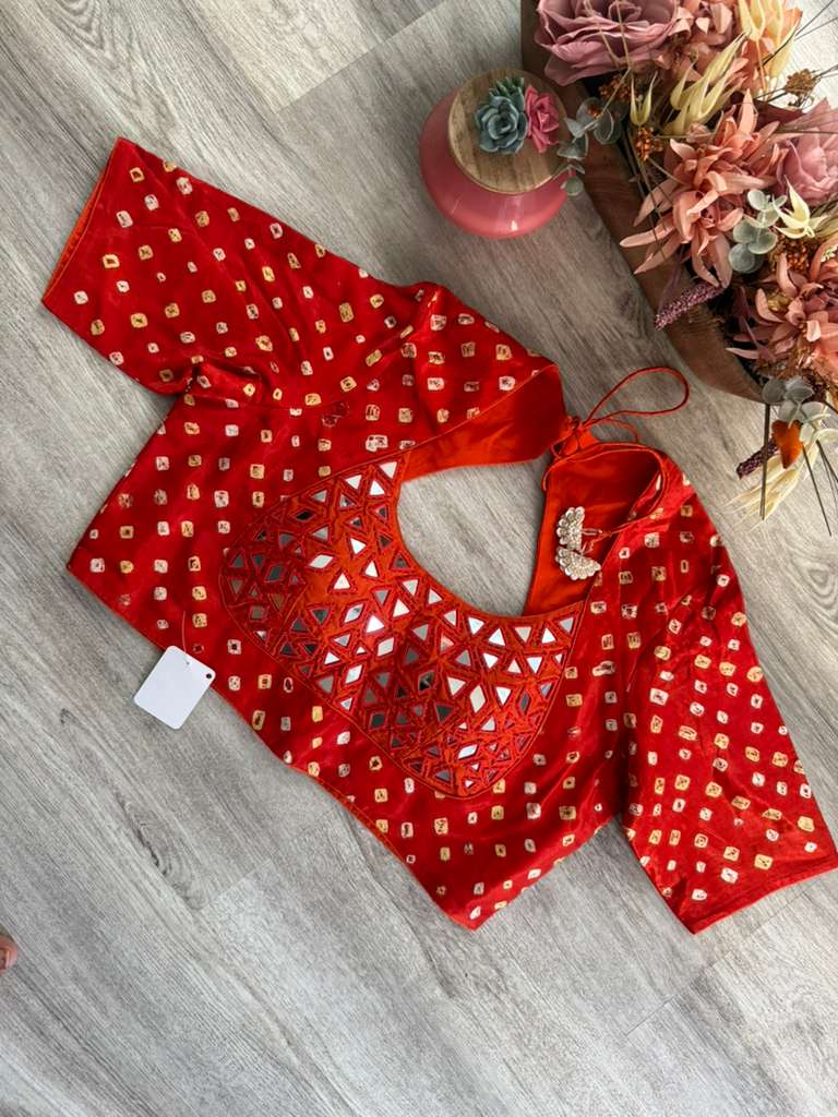 A red elbow-sleeve blouse with intricate bandhani and mirror work on Gaji silk fabric, laid flat on a gray wood-textured floor with decorative dried flowers in the background.