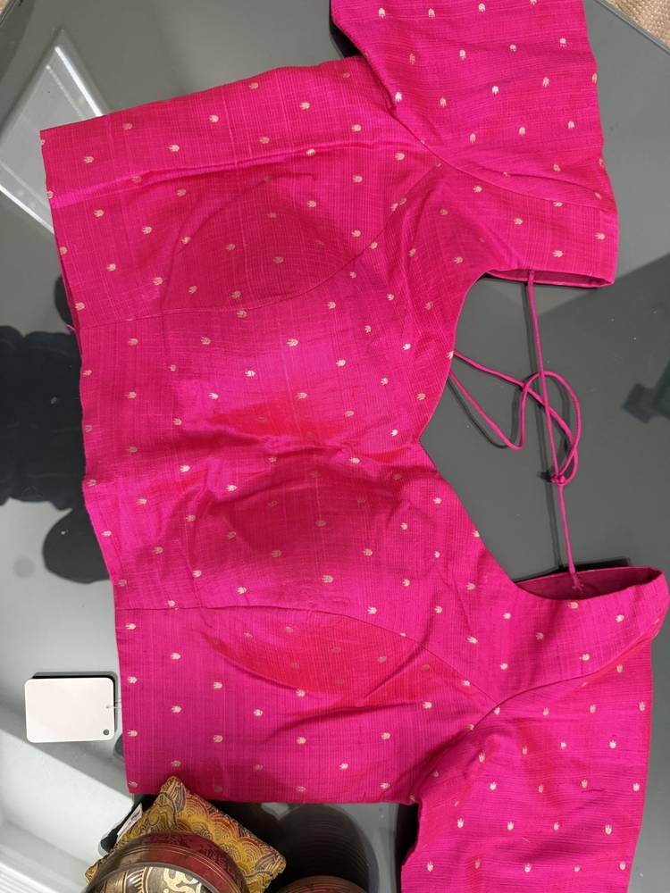 A bright pink cotton blouse with small white polka dots, laid flat on a dark reflective surface. The blouse has short sleeves, a round neckline, and a side tie detail.