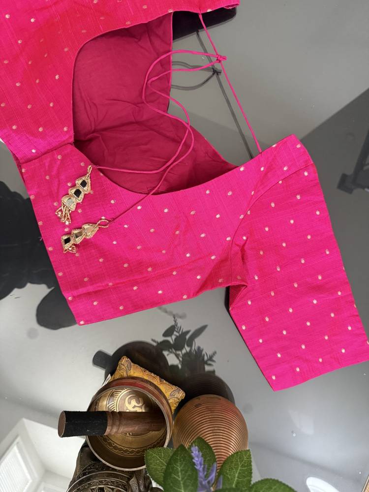 A bright pink cotton blouse with small white polka dots and gold embroidered details on the neckline, shown laid flat on a light gray surface with decorative props at the bottom.