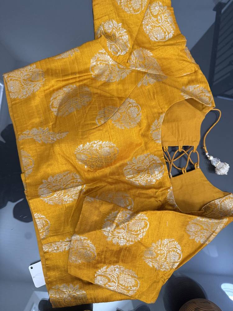 A mustard yellow saree with a silver floral brocade pattern, featuring a sleeveless blouse with side lacing and a tassel, laid flat on a gray surface.