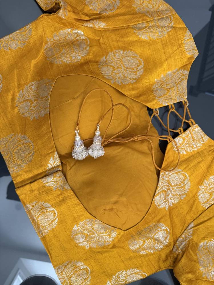 A mustard yellow and gold patterned traditional blouse set with matching skirt and a pair of silver tassel earrings, laid flat on a dark surface.
